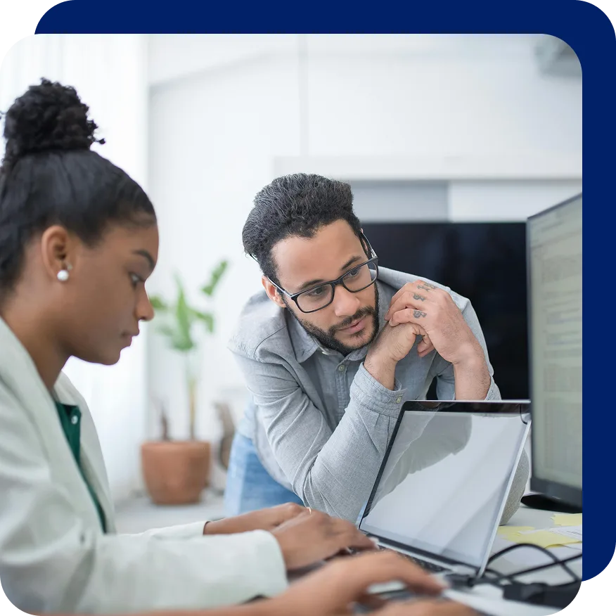 Two professionals collaborating in an office setting, with one person typing on a laptop and the other attentively observing and offering input, suggesting teamwork and business analysis