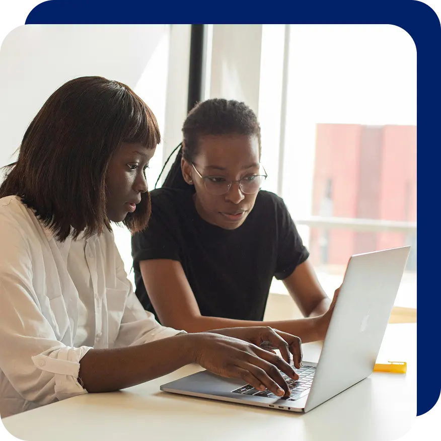 Two women collaborating on a laptop at a desk in a bright office setting