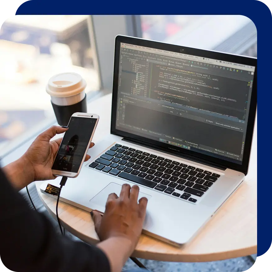 Person holding a smartphone connected to a laptop while testing mobile app code displayed on the screen