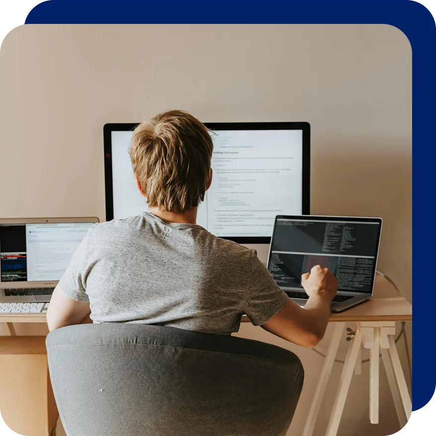 Rear view of a developer working at a desk with three screens displaying code and documentation, symbolizing multitasking and extended development capabilities