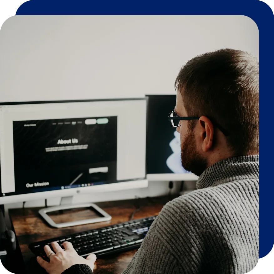 Man wearing glasses and a sweater working on a desktop computer, designing or editing an 'About Us' web page displayed on the screen