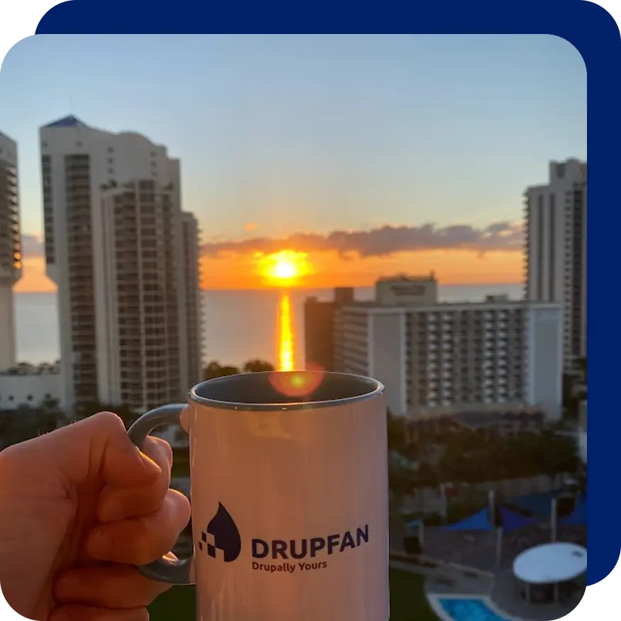 Hand holding a white mug in front of a city skyline at sunset, with high-rise buildings and the sun reflecting over the ocean.