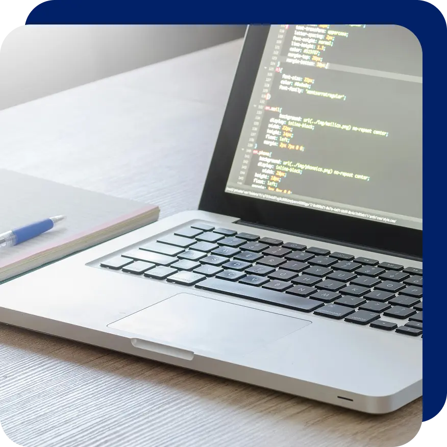 Laptop on a wooden desk displaying lines of CSS code, next to a notebook and pen, symbolizing coding, web development, or pay-per-click (PPC) ad campaign setup